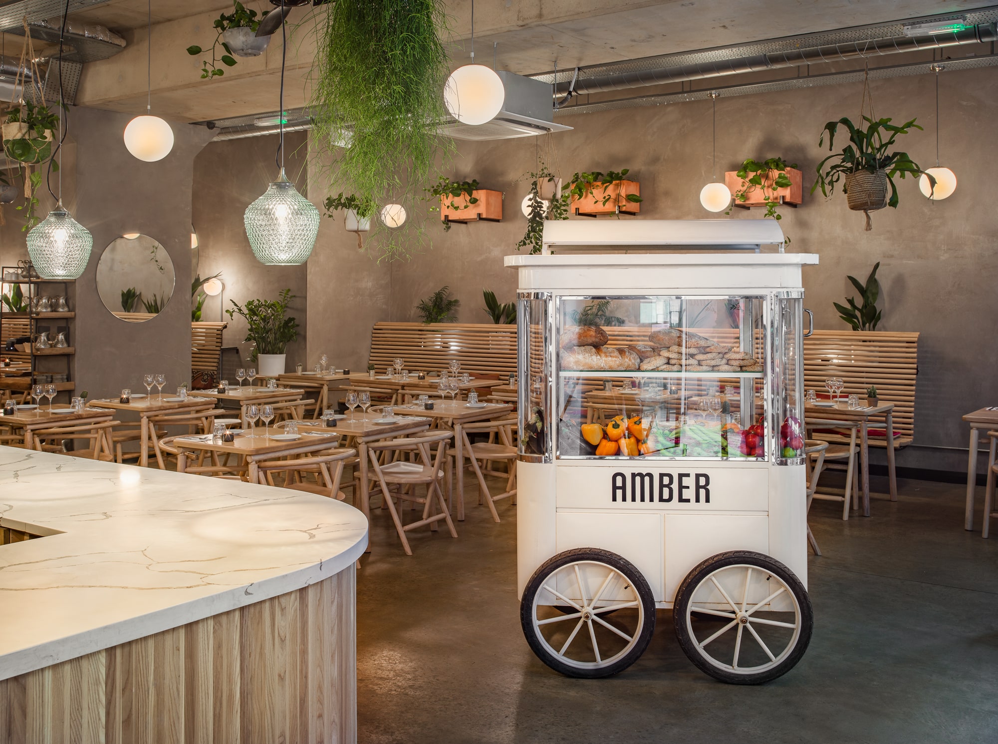 The bright and modern interior of Amber Restaurant in Aldgate East, featuring wooden furniture, hanging plants, and a stylish food cart with fresh produce and bread.
