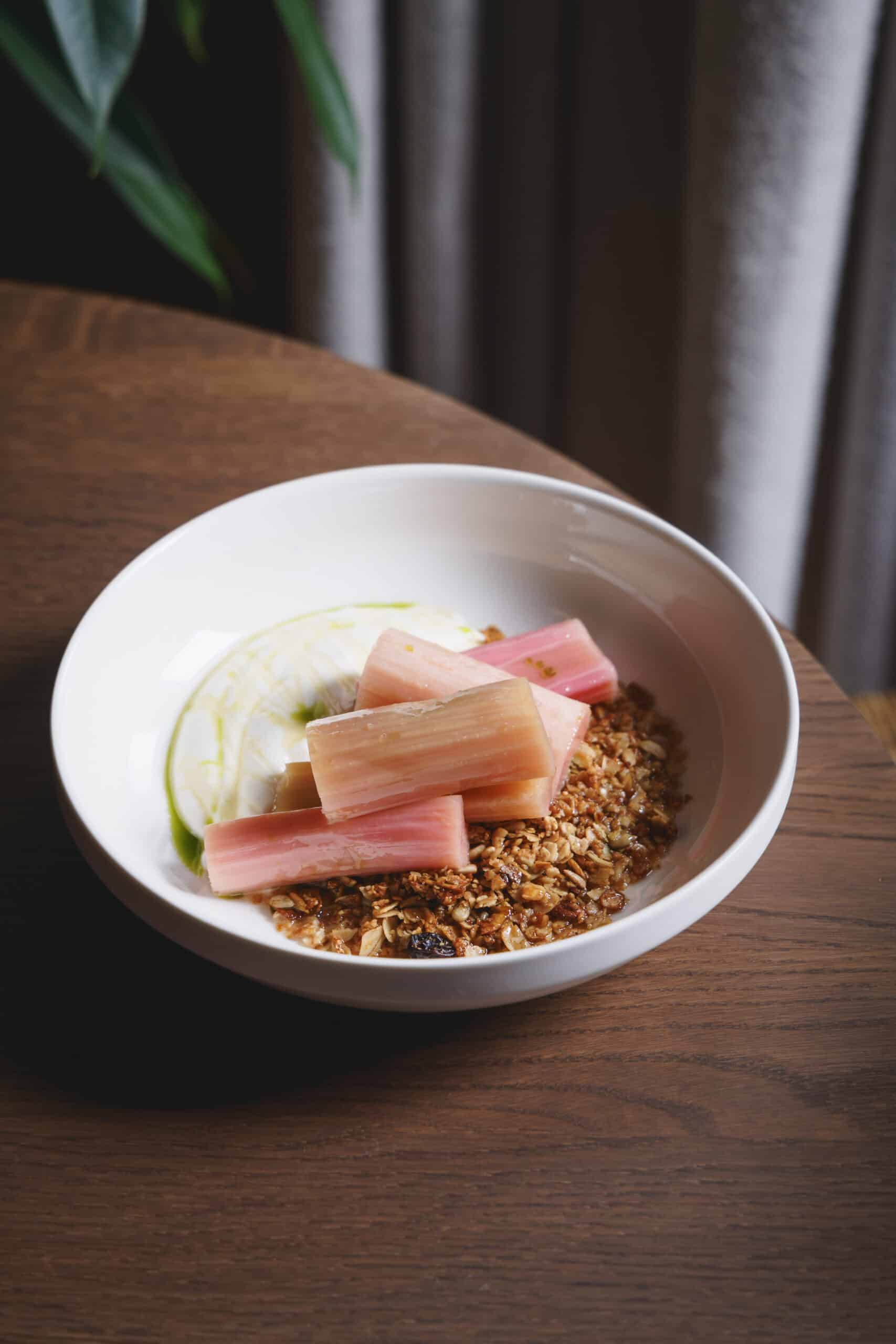 A breakfast bowl of poached rhubarb, granola, and yoghurt served at Ara Restaurant in Aldgate East, presented on a wooden table.