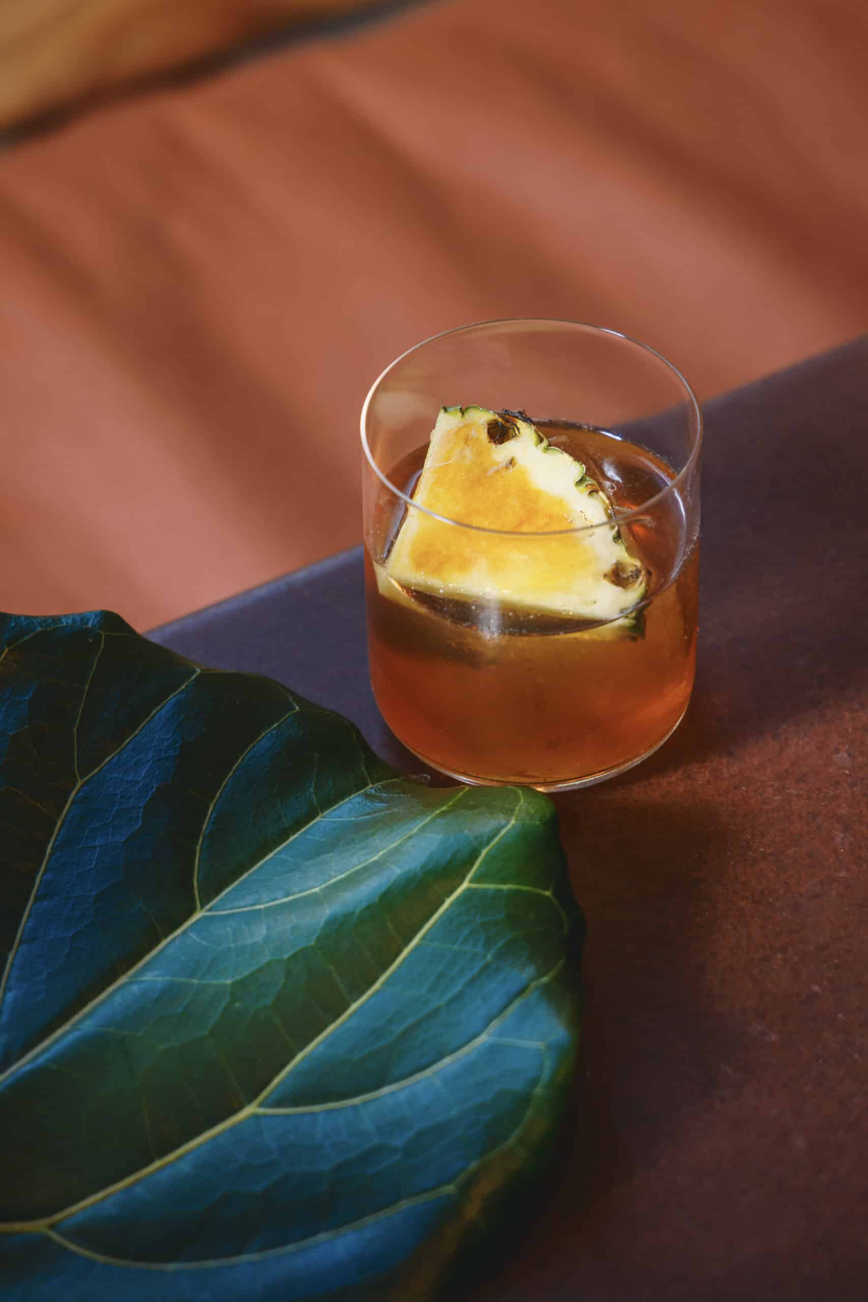 Banana peel rum cocktail with a pineapple garnish at Ara Restaurant, near Spitalfields and the City of London, served in a clear glass on a terracotta-toned surface.
