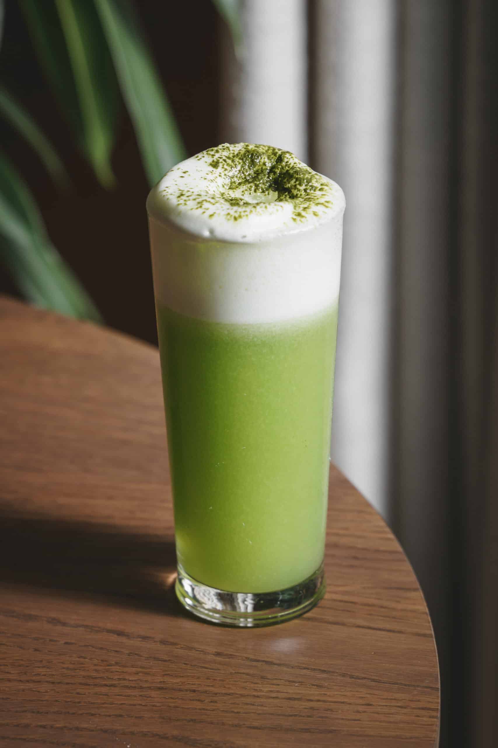 A bright green fizz cocktail with a frothy top, served at Ara Restaurant in the City of London near Spitalfields, presented in a tall glass on a wooden table.